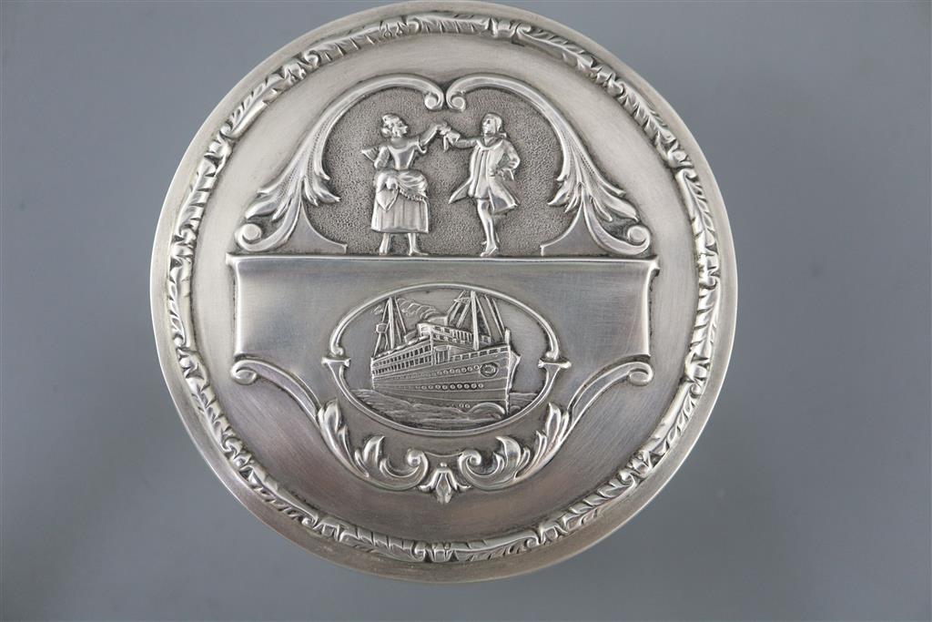 A George V silver circular box and hinged cover, embossed with a ship and dancing figures above,
